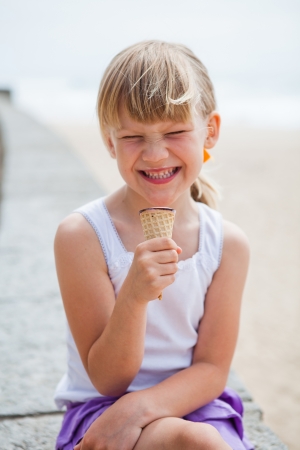 Young cute smiling girl holding ice cream while sitting on wall at beachの写真素材