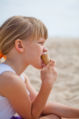 Young cute girl eating ice cream while sitting at beachの写真素材