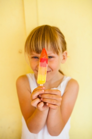 Smiling young girl holding up multicolored ice cream against yellow colored backgroundの写真素材