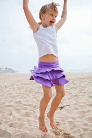 Happy barefoot young girl in purple skirt and white top jumping in sand on beachの写真素材