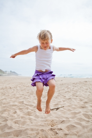 Happy barefoot young girl in purple skirt and white top jumping in sand on beachの写真素材