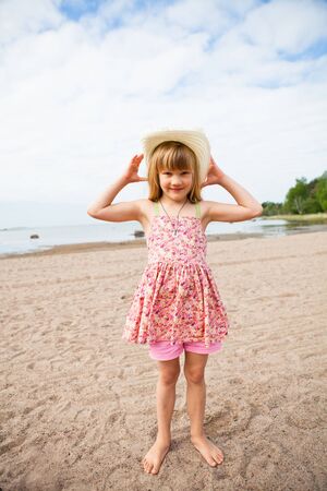 Smiling young barefoot girl wearing hat and pink patterned dress at beachの写真素材