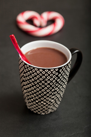 Closeup of mug of hot chocolate with red plastic spoon and hard candy heart with red swirls in backgroundの写真素材