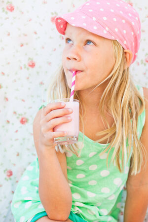 Cute young girl drinking milk fruit smoothie with striped straw from glass held in handの写真素材