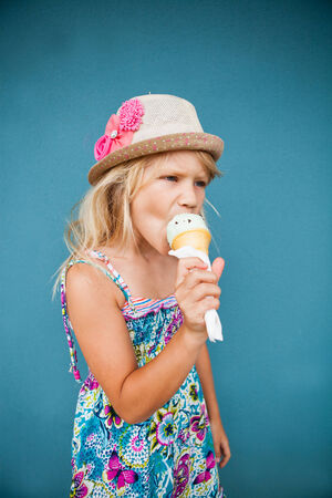 Cute young girl eating vanilla ice cream in cone outside against blue wall backgroundの写真素材