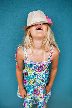Portrait of stylish cute young girl against blue wall background with hat covering eyes and funny expressionの写真素材