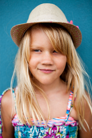 Portrait of cute smiling young girl wearing stylish hat outside against blue wall background and looking to sideの写真素材