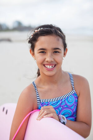 Portrait of cute smiling young girl holding pink surfboard at the beachの写真素材