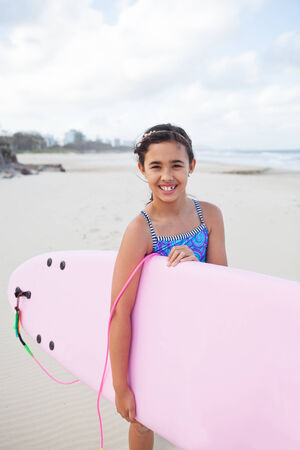 Cute smiling young girl holding pink surfboard at the beachの写真素材