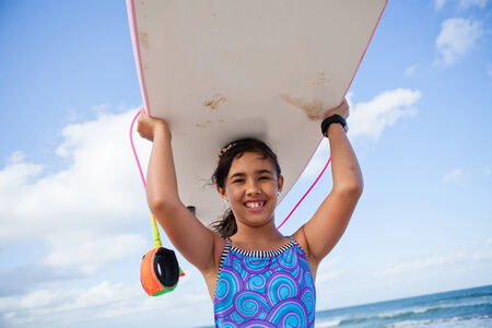 Cute smiling young girl holding pink surfboard on head at the beach with ocean and sky in backgroundの写真素材