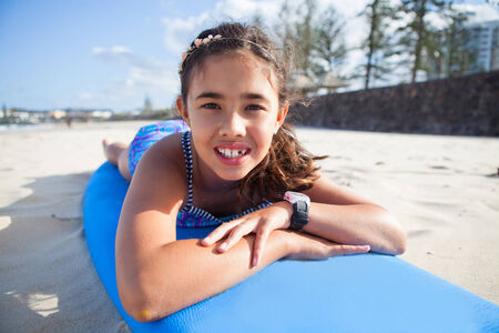 Closeup portrait of cute young girl lying on blue surfboard on the beachの写真素材