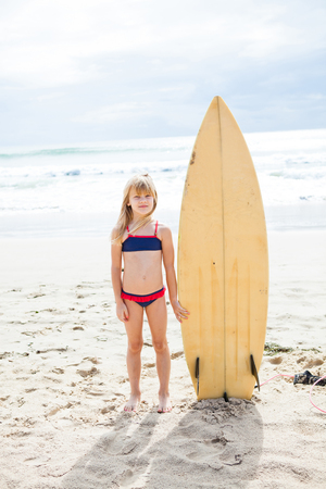 Cute young girl standing next to surfboard on beach with ocean in backgroundの写真素材