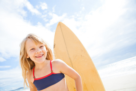 Portrait of happy smiling cute young girl with surfboard on beach with sunny sky backgroundの写真素材