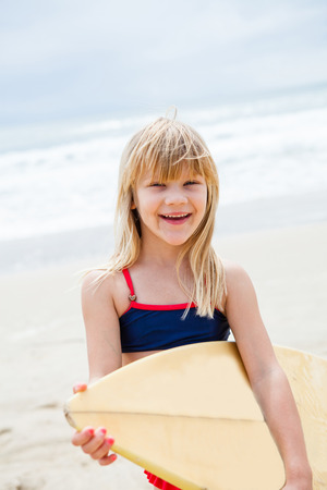 Happy smiling cute young girl with surfboard on beach with ocean in backgroundの写真素材