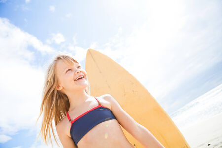 Portrait of happy smiling cute young girl with surfboard on beach with sunny sky backgroundの写真素材