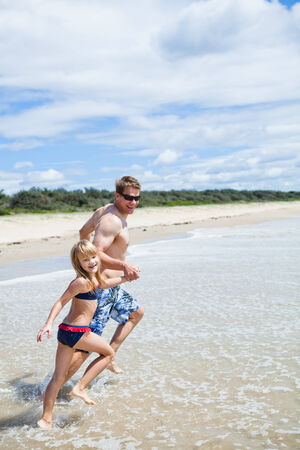 Happy smiling father and young daughter holding hands and running along beach in shallow waterの写真素材
