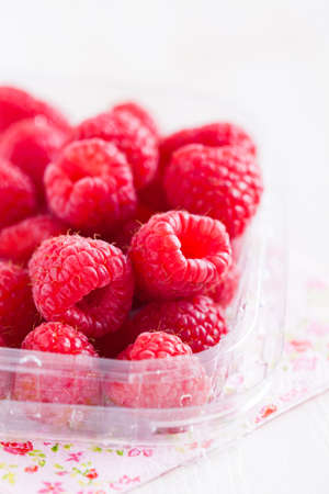 Closeup of bunch of fresh red raspberries in clear plastic container resting on decorative floral serviette on light surfaceの写真素材