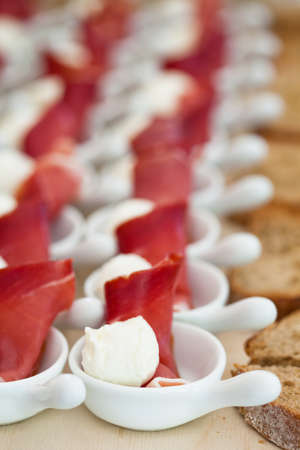 Closeup of prosciutto ham and ball shaped mozzarella cheese appetizers in rows of small white serving dishes and row of bread slicesの写真素材