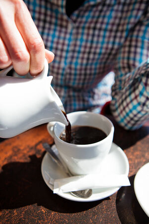 Closeup of pouring of black european style espresso based filter coffee into cup from jug by hand of woman at table at outdoor cafeの写真素材