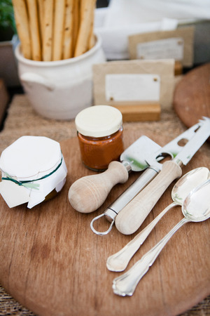 Closeup of various serving utensils and condiments in jars on wooden board with bread sticks in bowl in backgroundの写真素材