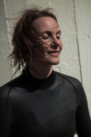 Attractive smiling brunette female swimmer wearing black wetsuit with wet hair and eyes closed and sunlight in faceの写真素材