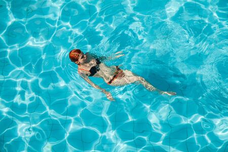 Young redhead woman in orange and black swimwear in swimming pool 