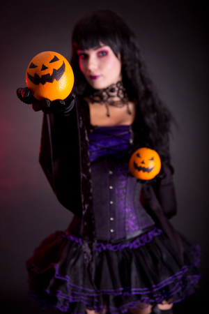 Beautiful witch in purple gothic Halloween costume with Jack lantern oranges, selective focus on fruits の写真素材