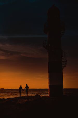 The silhouette of a couple and a huge lighthouse against the backdrop of the sea and the setting sun. Mexico.の写真素材