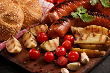 Grilled bbq sausages with vegetables, spices and bread on wooden cutting board backgroundの写真素材