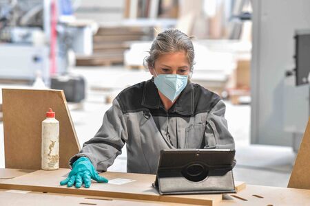 Woman reading technical informations on her tablet in a  industrial carpenter workshop and wearing protections against coronavirusの写真素材
