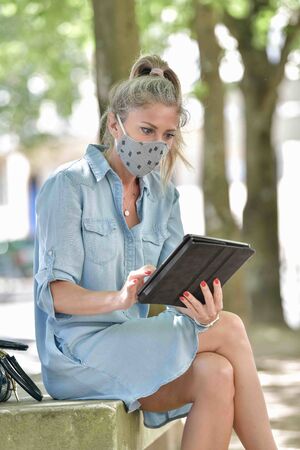 Woman doing a break outdoors  during the shutdown, reading  on her digital tablet while she is wearing a protective mask against the covid-19の写真素材