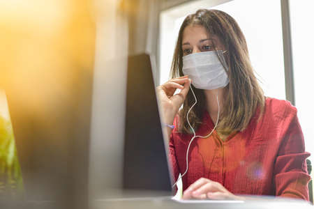 Young female woman wearing a protective mask while she is working at home and attending at a meeting on her computer during the lockdown due to the coronavirusの写真素材