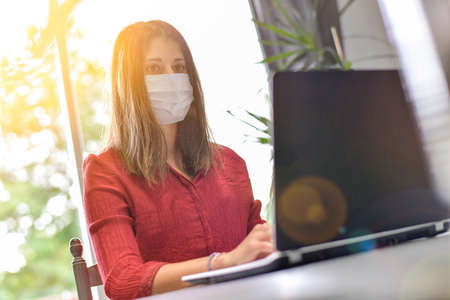 Young woman wearing a protective mask while she is working at home and attending at a meeting on her computer during the lockdown due to the covidの写真素材