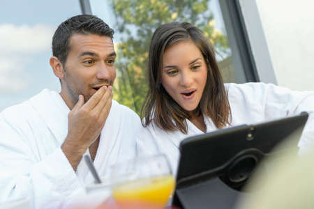 Happy young couple in bathrobes having breakfast outdoors while they are on doing a video callの写真素材