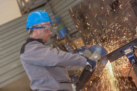 Concentrated male worker wearing protective cloths cutting a metal bar with grinding machineの写真素材