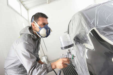 Car body worker paints a car in the paint booth with a spraying paint and a protective maskの写真素材