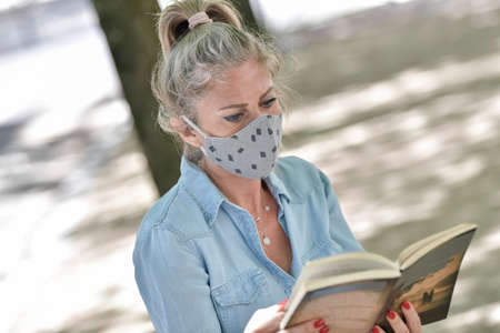 Woman doing a break outdoors during the shutdown, reading a book while she is wearing a protective mask against the covid-19の写真素材