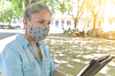 Woman doing a break outdoors during the confinement, reading on her digital tablet while she is wearing a protective mask against the coronavirusの写真素材