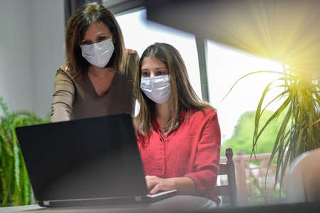 Mother and daughter wearing protective mask and attending online video learning lesson during lockdown due to coronavirusの写真素材