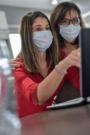 Mother and daughter wearing protective mask and browsing and shopping on the web at home on their computer during lockdown due to covid-19の写真素材