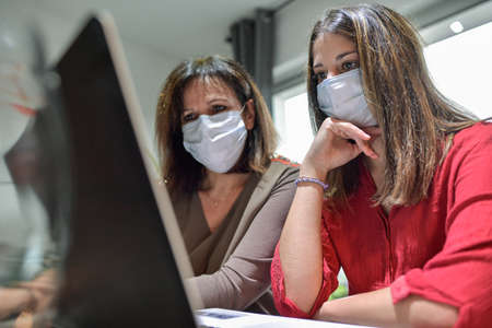 Mother and daughter wearing protective mask and attending online video learning lesson during lockdown due to coronavirusの写真素材