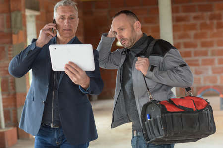 Mature architect on the phone is holding a tablet while standing next to a worker inside of a siteの写真素材
