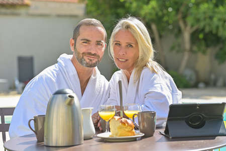 Portrait of a mature couple having breakfast in bathrobe outside in front of their swimming poolの写真素材