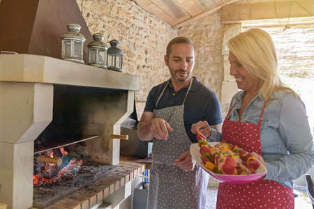 Relaxed couple cooking skewers of beef on the barbecue in a fireplace in a external kitchenの写真素材