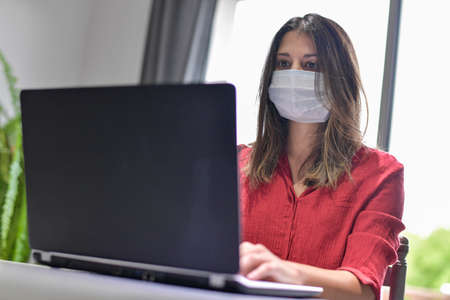 Young female woman wearing a protective mask while she is working at home and attending at a meeting on her computer during the lockdown due to the coronavirusの写真素材