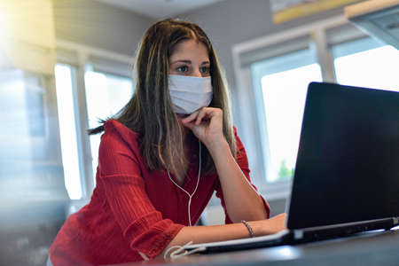 Young female worker wearing a protective mask while she is at home and attending at a meeting on her computer during the lockdown due to the coronavirusの写真素材