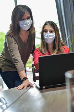 Mother and daughter wearing protective mask and attending online video learning lesson during lockdown due to coronavirusの写真素材