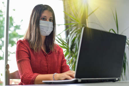 Young woman wearing a protective mask while she is working at home and attending at a meeting on her computer during the lockdown due to the covidの写真素材