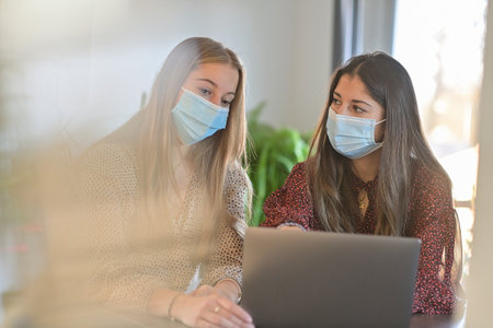 Twin sisters wearing a protective mask while they are working at home and attending at a meeting on their laptop during the lockdownの写真素材