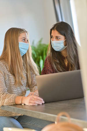 Twin sisters wearing a protective mask while they are working at home and attending at a meeting on their laptop during the lockdownの写真素材